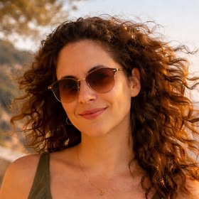 Christine, wearing tinted aviator sunglasses and an olive top, outdoors in warm evening light.
