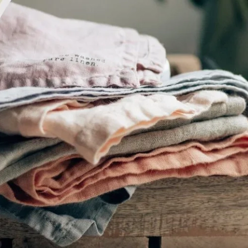A stack of folded linen garments in mauve, blue-grey, cream, sage, and coral on weathered wood.