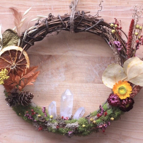 A twig wreath with moss, clear quartz points, dried orange, pinecones, and dried flowers, hanging by a jute cord.